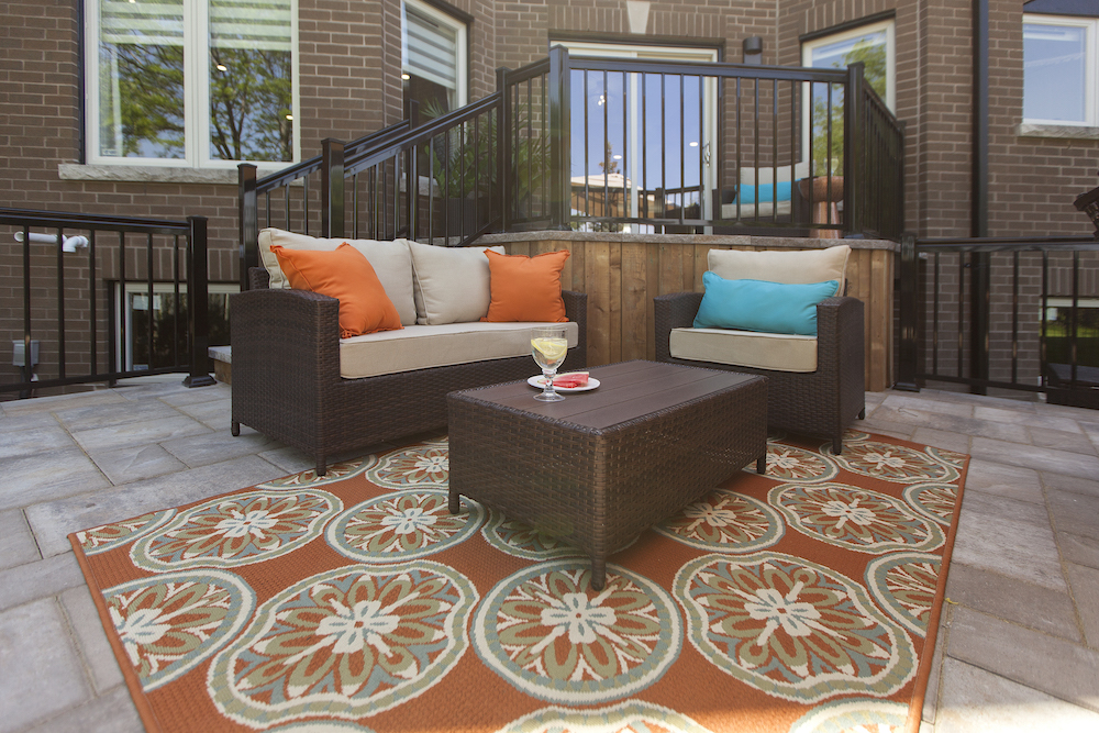 Large deck at the back of a brick suburban house decked out with brown patio furniture, orange and teal pillows and orange weather resistant rug