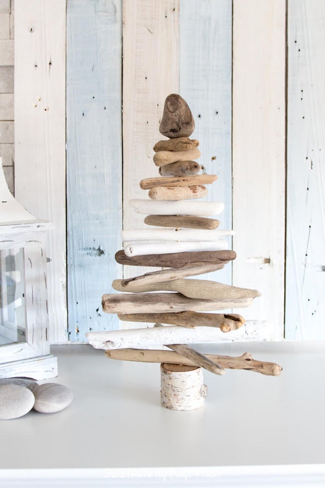 A tabletop tree made from salvaged driftwood sits on a white countertop beside a white lamp and three white stones against a white and blue shiplap wall