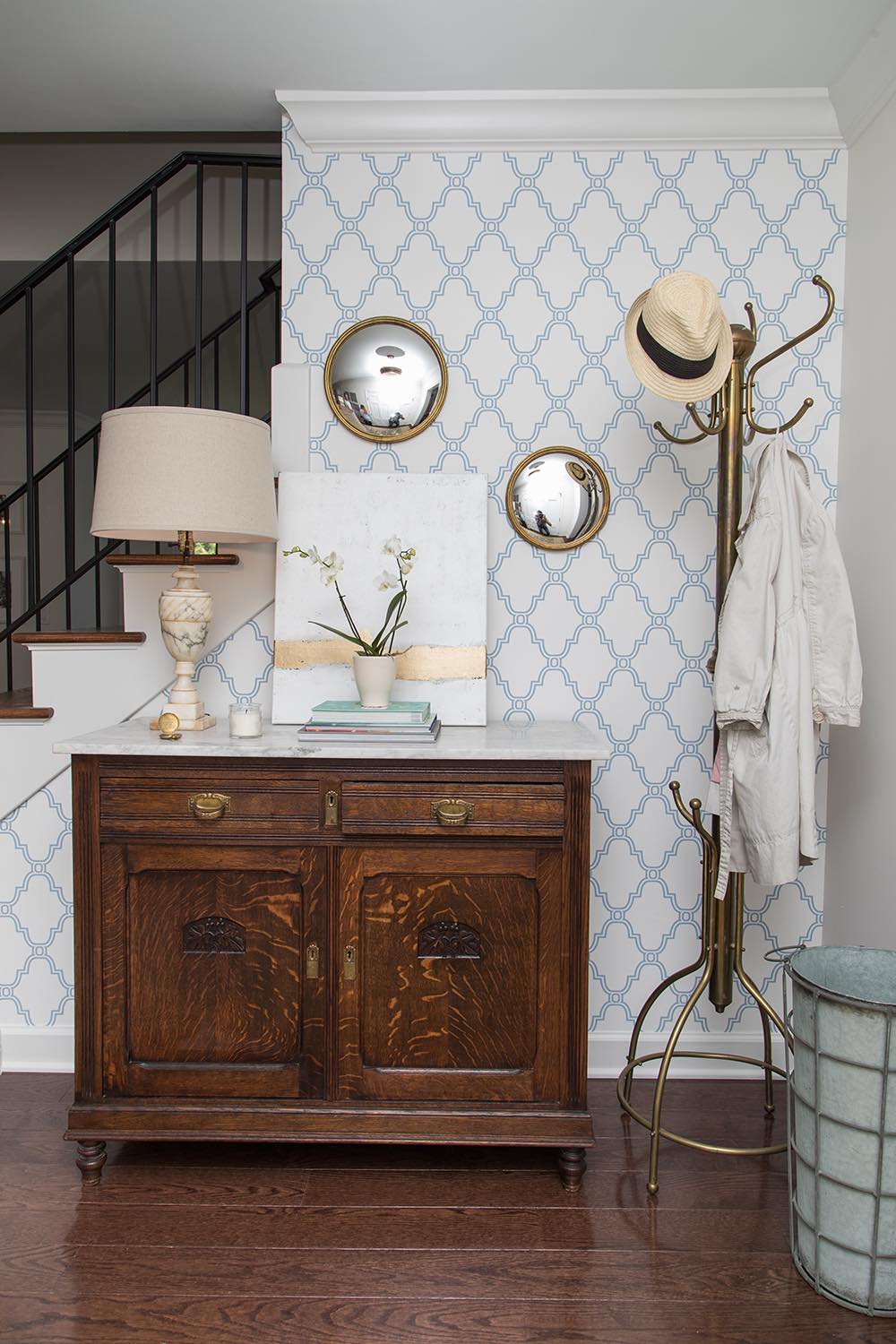Beautiful entryway set up at the base of a set of stairs with an antique wooden floor cabinet topped with a classic lamp, a stack of books, a potted orchid and a white and gold art canvas, in front of a wall covered in blue and white geometric wallpaper and two circular convex mirrors, and a coat rack with a straw hat and white coat hanging of it