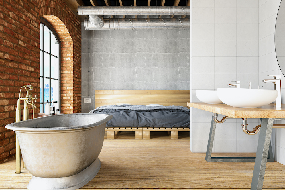 Open concept loft with a brick wall and large windows, a large metal soaker bathtub with brass hardware sitting in front of a two white vessel sinks wooden vanity and wooden bed with grey sheets