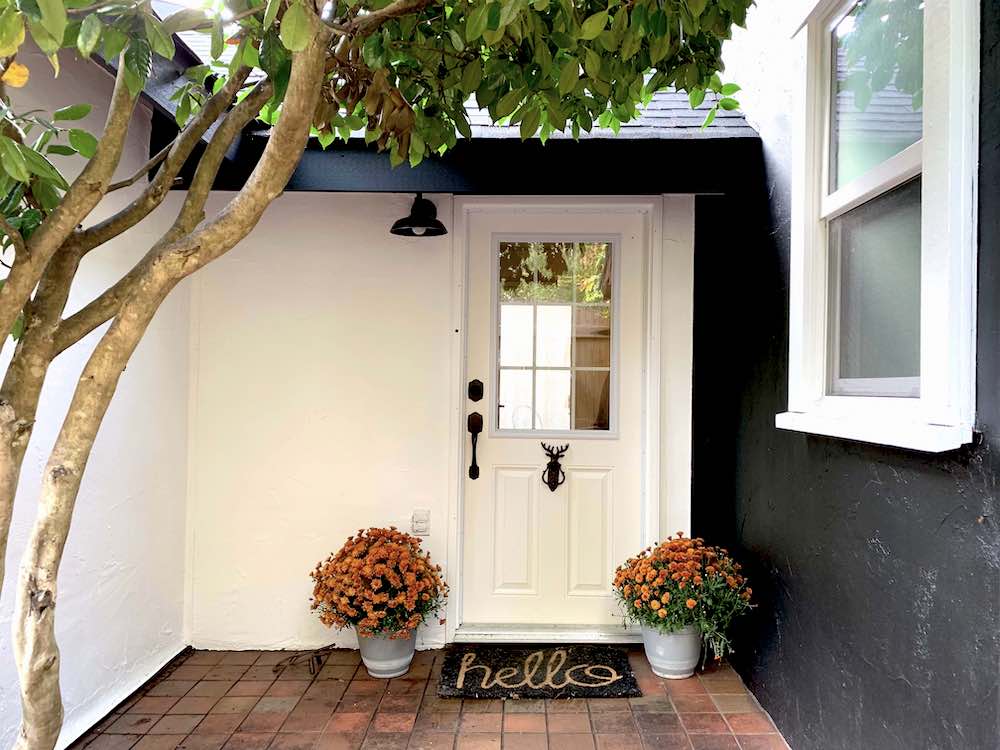 Entryway to a cottage with a white glass paneled door, a camellia tree, two grey pots of orange mums, a “hello” welcome mat, and exterior walls painted in BEHR Black Smoke PPH-34 and Ultra Pure White 1850