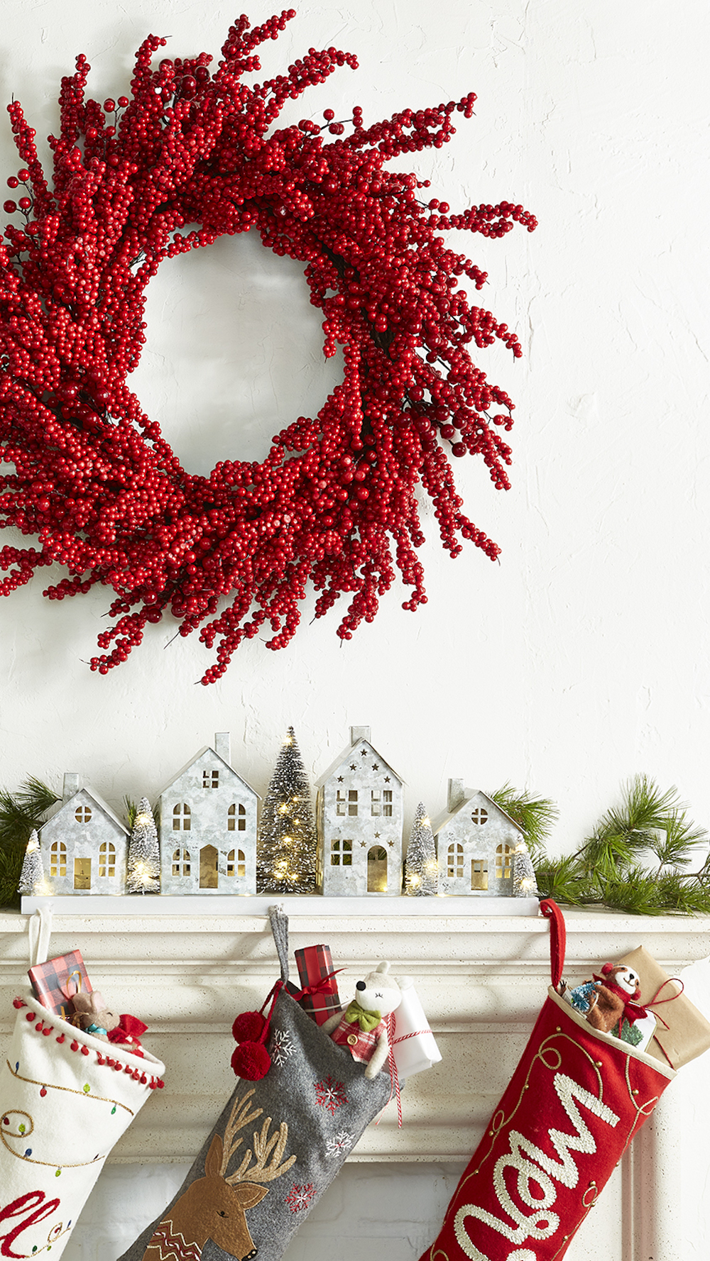 Christmas decoration scene featuring items from HomeSense with metal light up houses on a white mantle, a large red holly wreath hanging on the wall above it, and three stuffed stockings hanging down