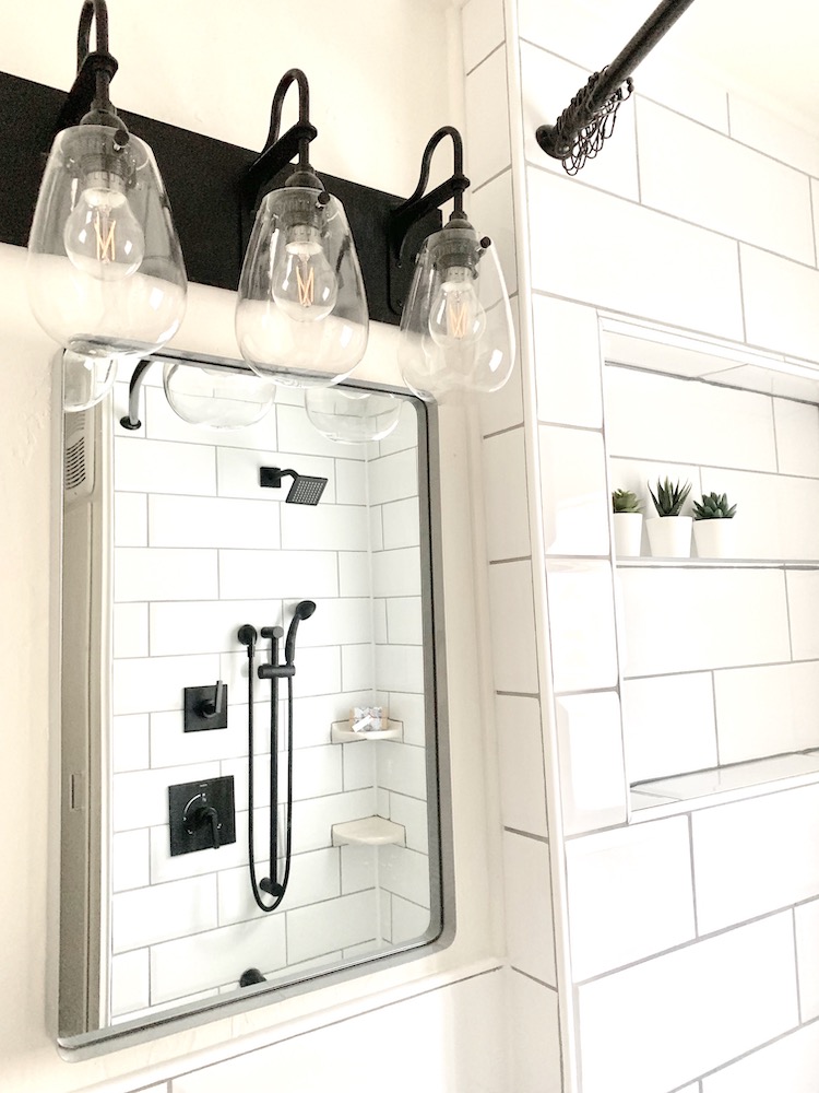Beautiful modern bathroom with large white shower tiles, grey mirror, black vanity lights, and a shower shelf with three potted faux succulents