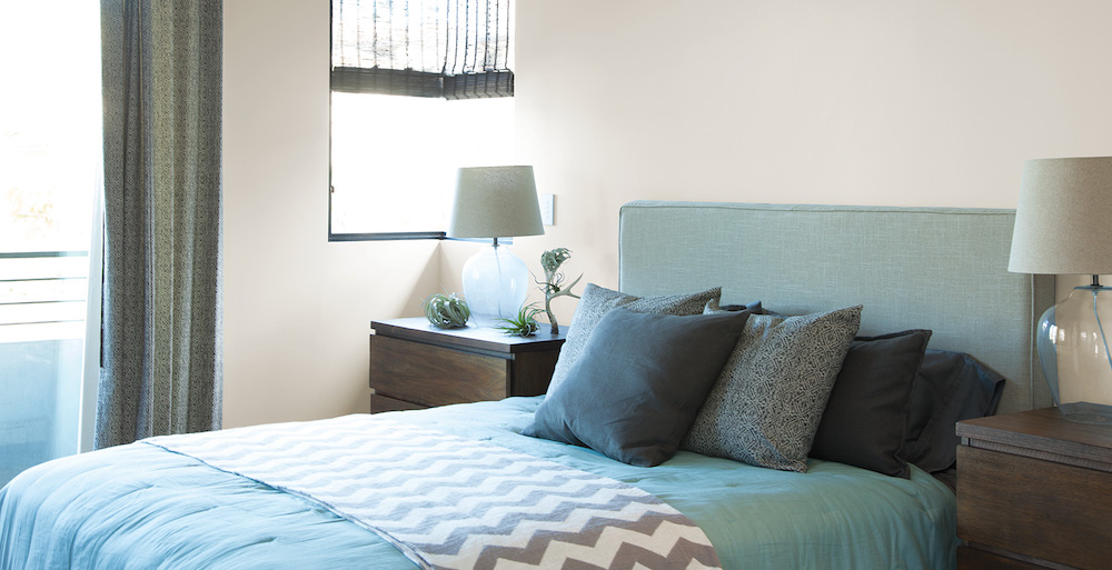 Simple modern bedroom with off white walls painted in BEHR Off White 73, a grey upholstered bed with a blue bedspread and a pile of grey pillows, and two dark wood bedside tables with a glass lamp on each.