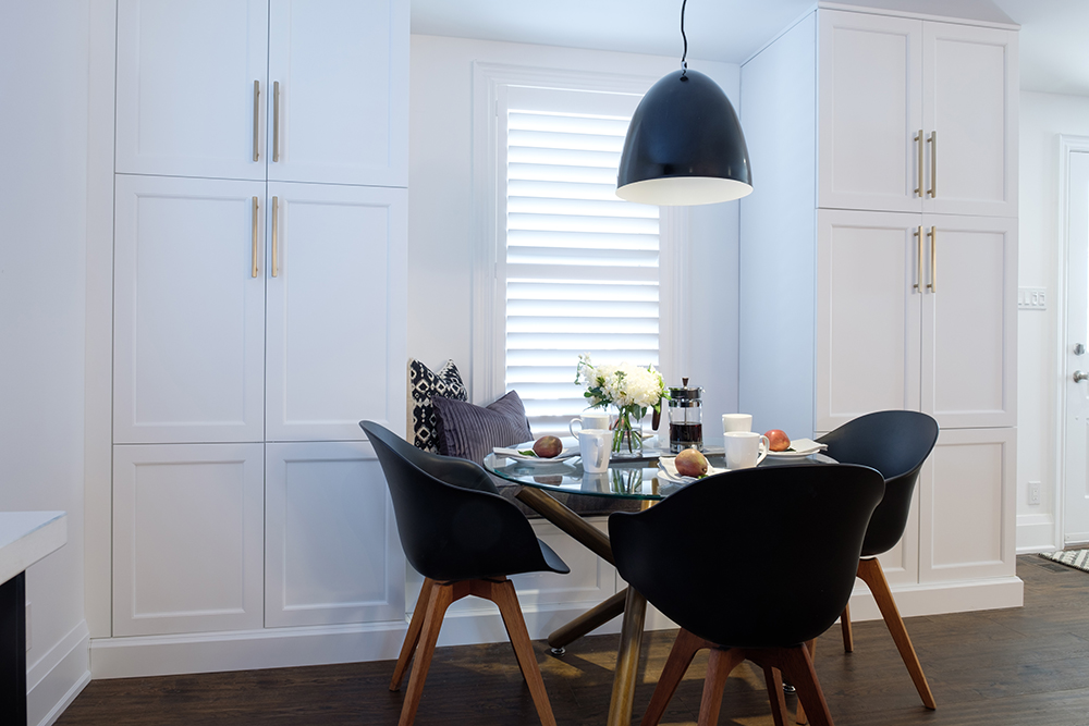 Chic black and teak mid century modern dining table and three chairs set in a white breakfast nook with large black pendant lamp overheard
