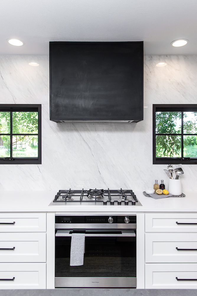 Chic modern kitchen with white cabinets with black pulls, a high end stovetop and oven, two black framed windows, and a black square oven hood