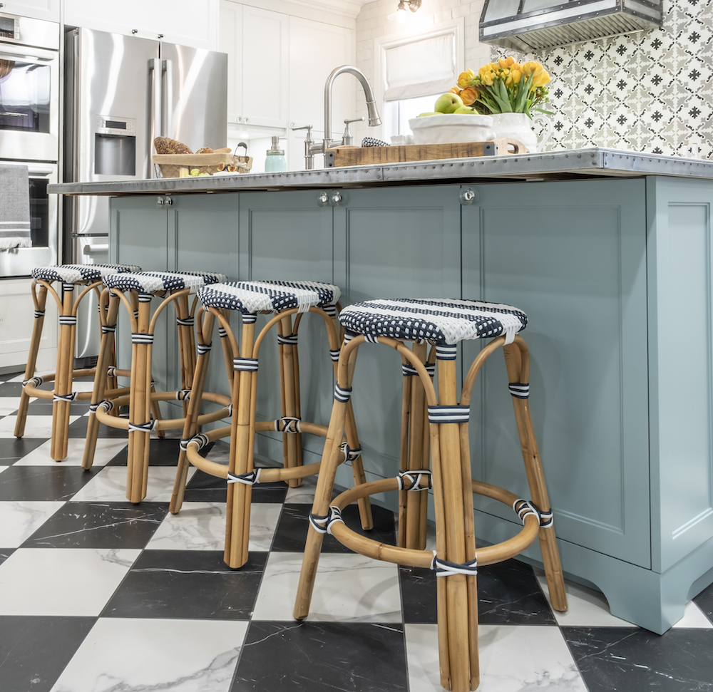 Four rattan chairs stand by a blue kitchen island on a black and white tiled floor