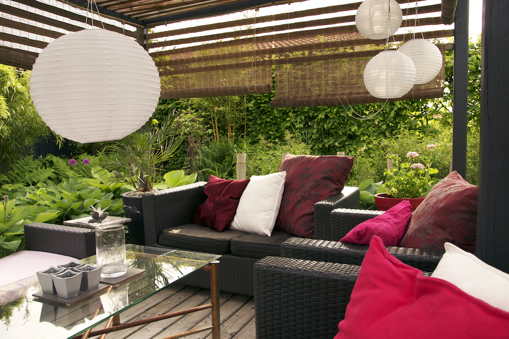 A gorgeous garden filled with plants and flower growing up around a stunning wood pergola over a large wicker outdoor couch covered with a red and white cushions and a glass topped coffee table