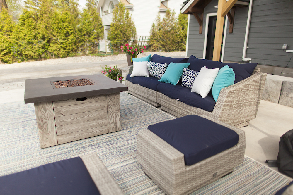 Grey patio furniture decorated with blue and white pillows places around a gas fire pit