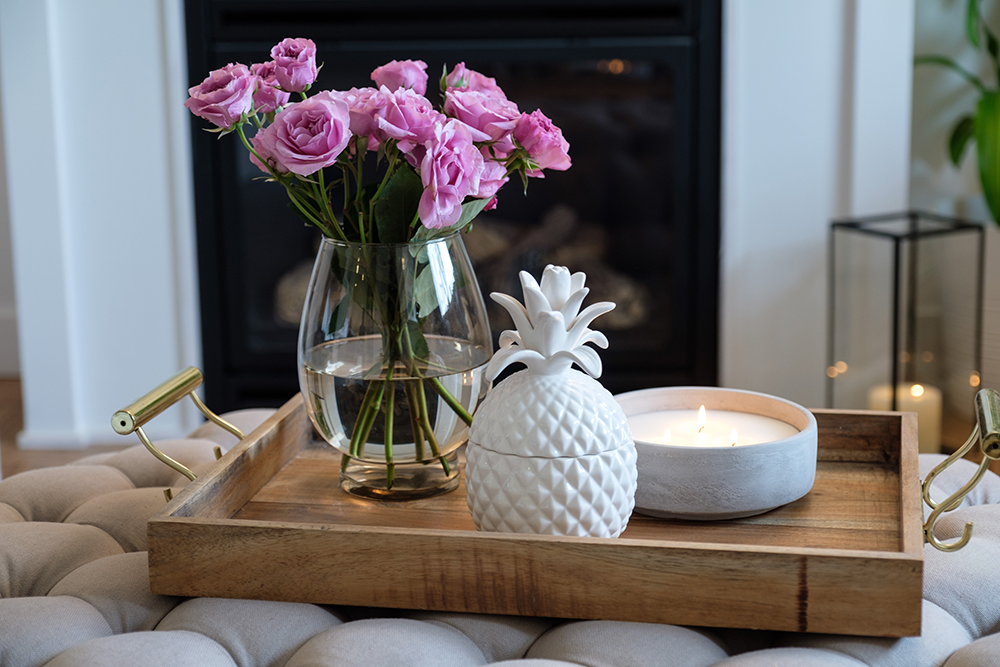 A wooden tray with pink roses, a candle and a pineapple jar sit on top of a tufted ottoman