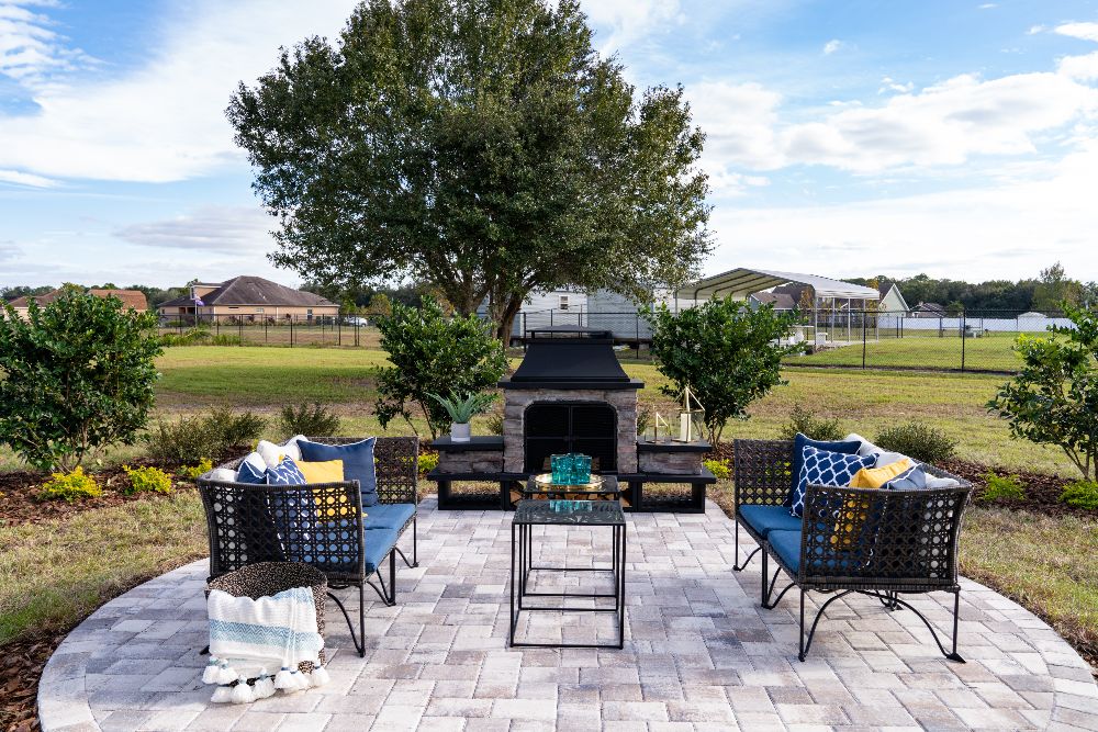 An outdoor entertaining area with stone pavers, two outdoor love seats around glass-topped tables, and a large outdoor fireplace.