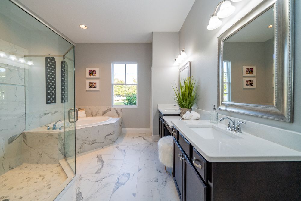 A main suite bathroom with large walk-in shower, separate soaker tub, and dual sinks with mirrors separated by a vanity table and plush ottoman.