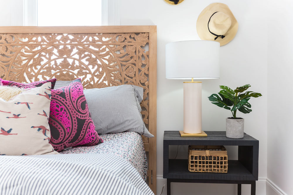 Masters of Flip bohemian Victorian house bedroom with ornately carved headboard, throw pillows, bedside table with lamp and a plant and a hat hanging on the wall