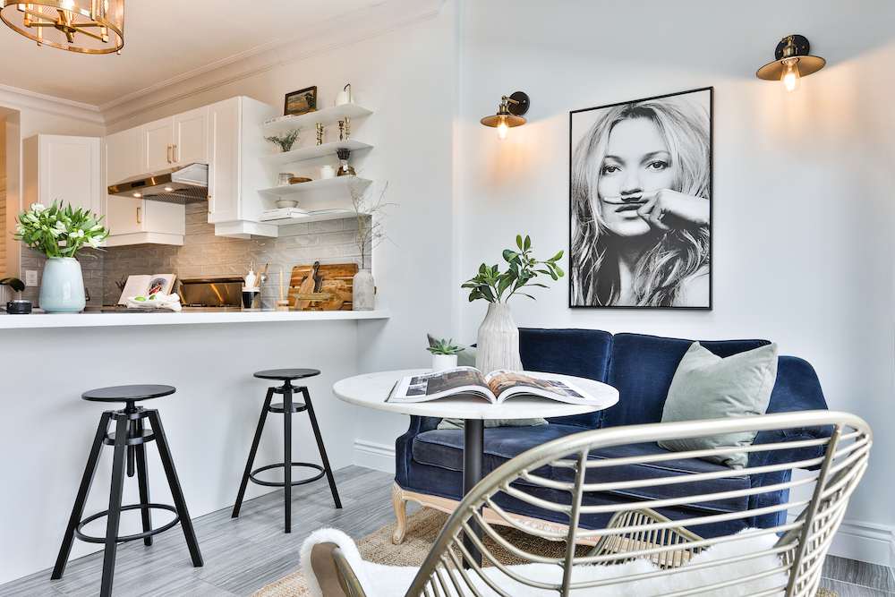 Chic white eating nook designed and styled by The Property Stylist Inc. with a blue couch and a white round table, with two wall scones and Kate Moss portrait in front of an all-white kitchen island with two black stools