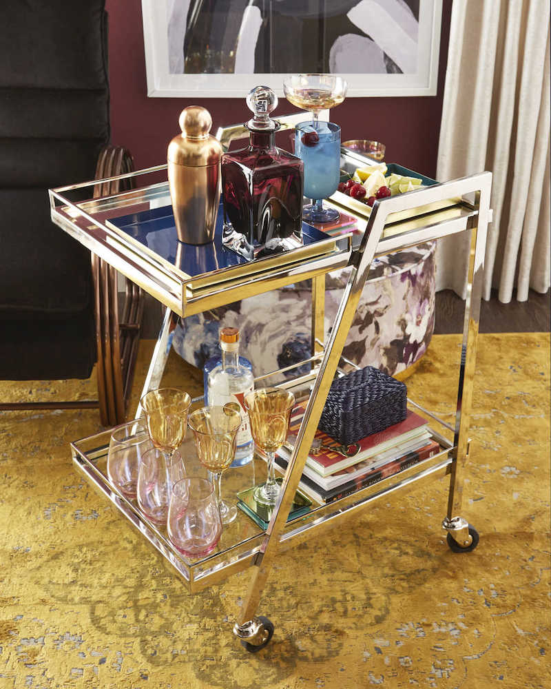 Small modern rectangular chrome bar cart with two mirrored shelves filled with cocktail making tools, glassware and bottles