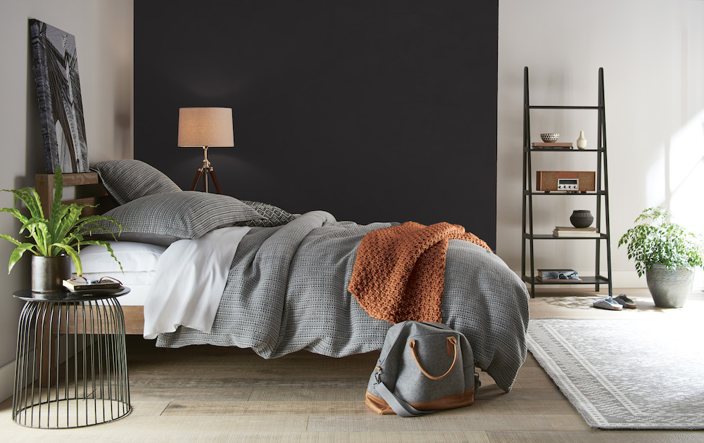 A luxurious bedroom with a grey upholstered bed, end of bed bench, grey pillows and comforter, a black bedside table with a grey lamp, and walls painted in BEHR Smoky White BWC-13 and Broadway PPU18-20 and accents painted in BEHR Maple Glaze PPU3-16 Accent: Maple Glaze PPU3-16
