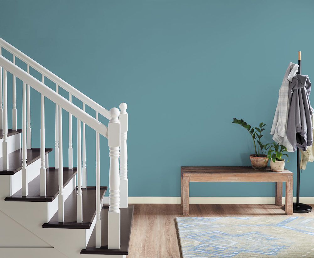 White staircase and baseboards painted in BEHR Polar Bear 75, light wood flooring, and area rug with subtle pattern, a wood bench topped with potted plants near a wall painted blue in BEHR Voyage PPU13-07