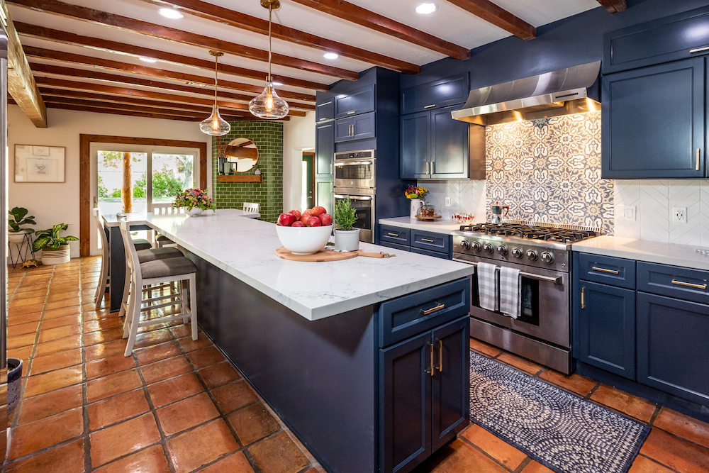 Charming large kitchen with navy blue cabinets, geometric blue and grey tile backsplash, stainless steel appliances, white quartz island surrounded by white chairs and exposed beams on the ceiling