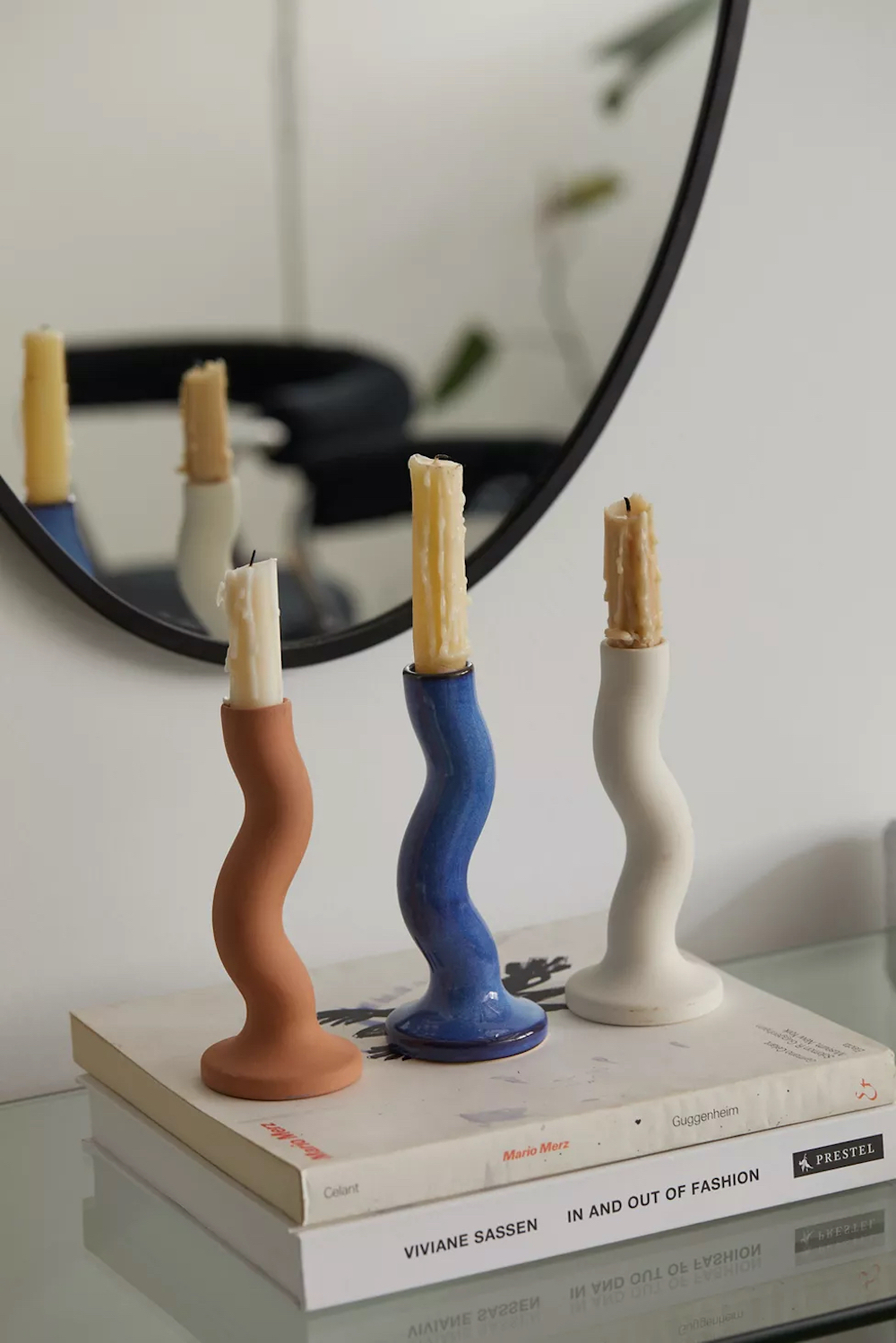 Three wavy taper candle holders sculpted from ceramic in a glazed finish from at Urban Outfitters with three half burnt candles on top of a stack of two books on the console table with a round black framed mirror behind