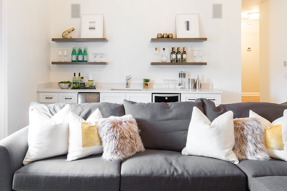 Fun modern bonus room with a large grey couch covered in white, gold and faux fur throw pillows, a white mini bar with a fridge, dishwasher and four floating shelves