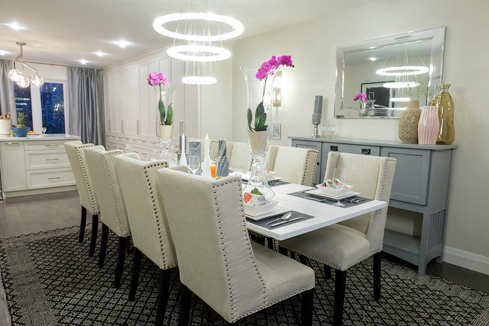 A chic sparkling tiered chandelier hangs about a white dining room table and white upholstered chairs