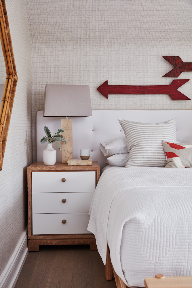 Chic bedroom with textured cream coloured wallpaper, a wooden bedside table with a lamp, and two red wooden arrows hanging on the wall