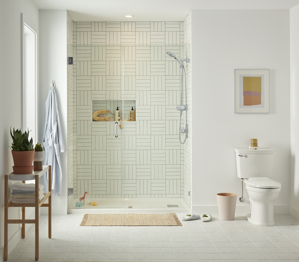 Chic white bathroom with white square floor tiles, a towel stand with two potted plants, a white toilet to the right, and a walk-in shower with woven white tile and a Spectra Filtered 4-Spray Hand Shower Rail System from American Standard found at Splashes Bath & Kitchen