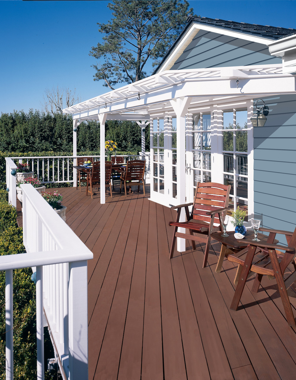An exterior deck painted in at the back of a blue house painted in BEHR Rainy Season MQ5-27 with white trim and pergola painted in BEHR Ultra Pure White with two wooden tables surrounded by deck chairs