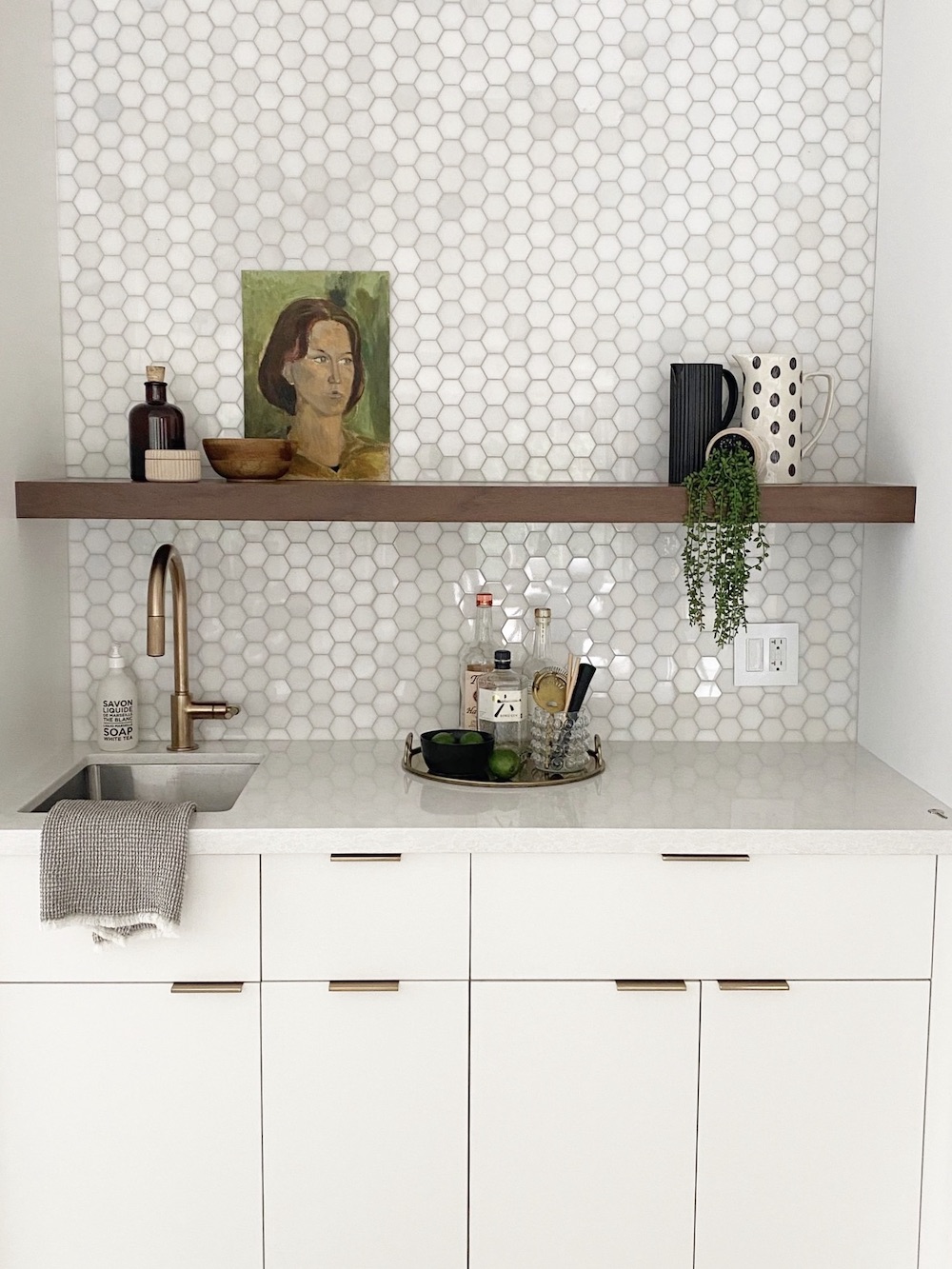 Small kitchen bar area designed and styled by The Property Stylist Inc. with white countertops, white hexagon tile backsplash, a wooden shelf, and brass accessories