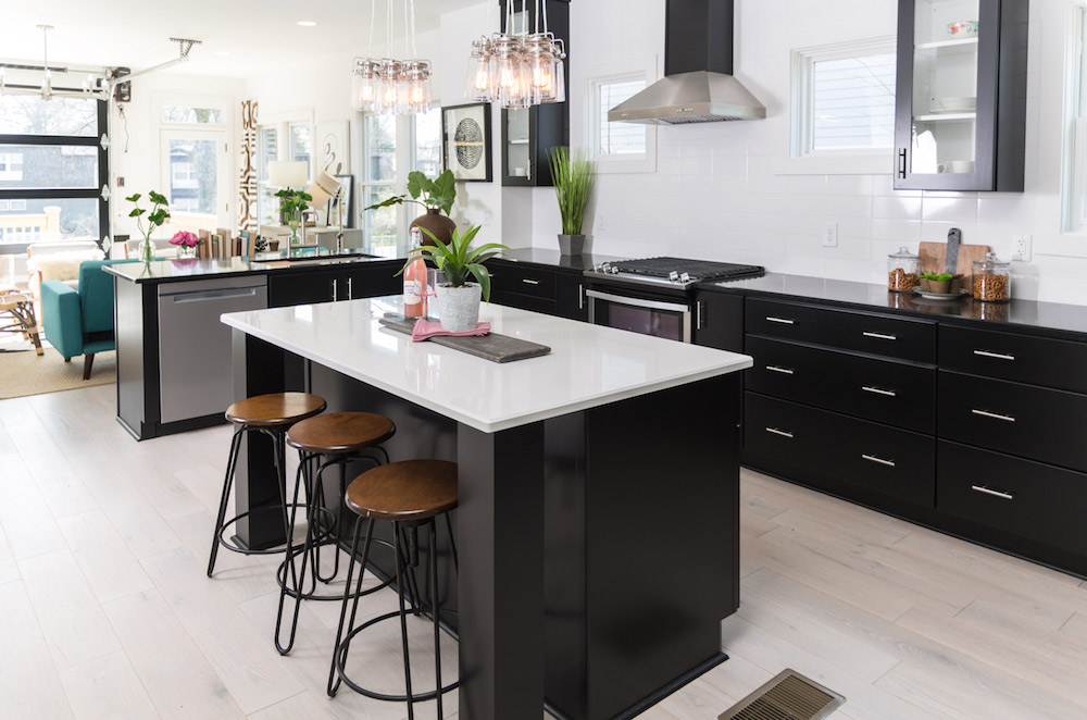 Chic modern kitchen with light wood floors, a large centre island with three barstools and a white countertop, black cabinets, black counters, black and stainless-steel appliances, and two groups of glass pendant lights as featured on Master of Flip on HGTV Canada