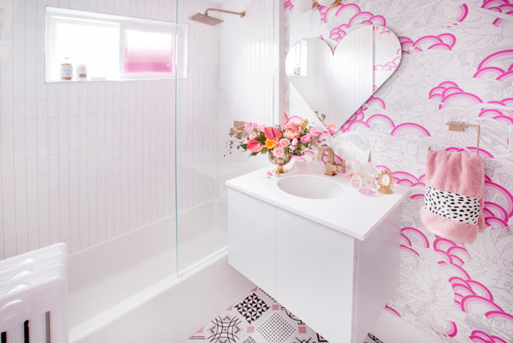 Bright white and pink bathroom designed by Tiffany for HGTV Canada’s Home to Win! with white, black and pink tiled floors, white, grey and pink patterned wallpaper behind a floating white vanity and sink with a heart-shaped mirror, and a white shower tub combo with a square brass rain shower head