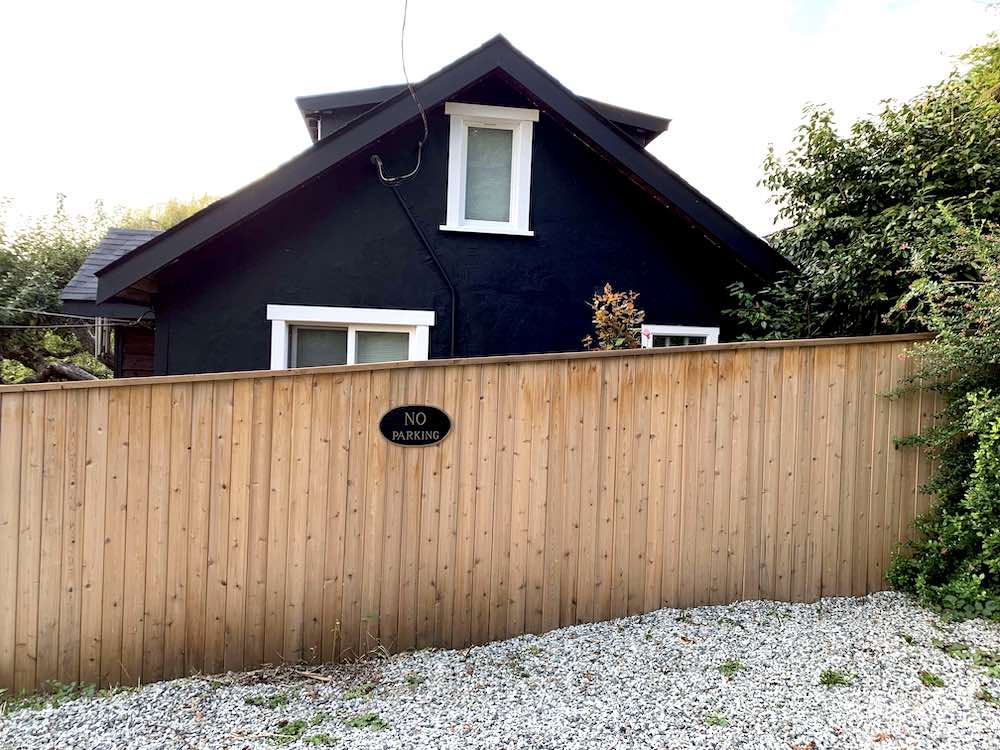 A gravel parking spot and a tall cedar plank fence in front of the exterior of a black and white cottage painted with BEHR Black Smoke PPH-34 and Ultra Pure White 1850