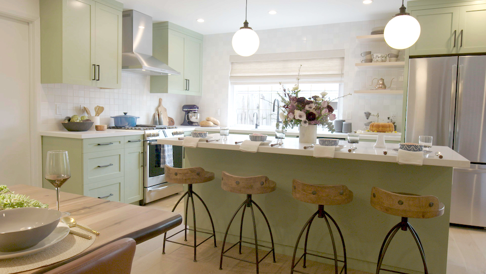 A newly renovated traditional kitchen with mint green painted cabinets with black hardware, a large kitchen island with four barstools, white countertops, white tiled backsplash and stainless steel appliances designed by Jonathan and Drew Scott for HGTV’s Property Brothers: Forever Home