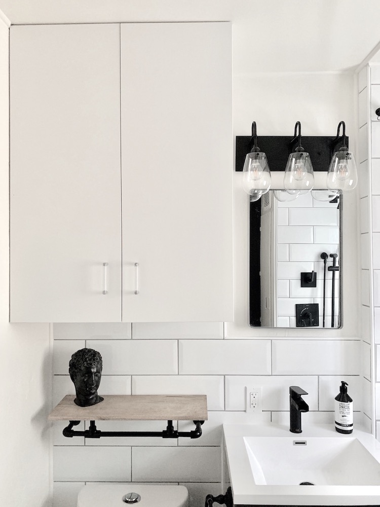 Beautiful modern bathroom with large white wall tiles, grey mirror, black vanity lights, white sink with a black faucet, large wall cabinet, and a floating industrial shelf with a black bust
