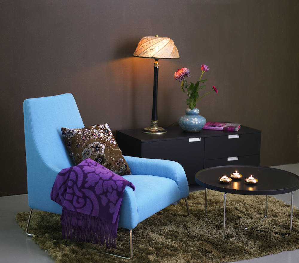 Blue armchair in a living room with a brown rug, small round coffee table, brown walls, and a black side table with a lamp and blue vase with flowers