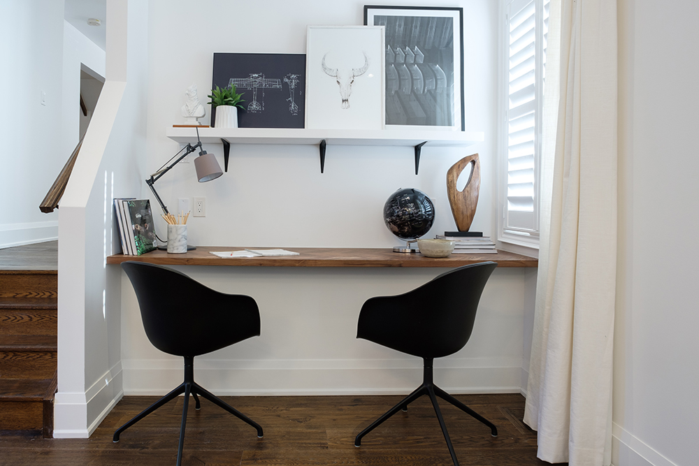Modern at home office area with two black bucket chairs, a wood desktop, and a white shelf with black brackets holding three pieces of framed artwork