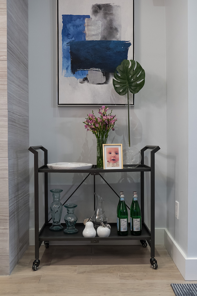 A blue painting hangs over a black bar cart stacked with vases, flowers and water bottles