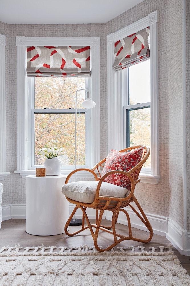 A bay window sitting area with a rattan armchair, white shagged rug, cream coloured textured wallpaper and roman blinds