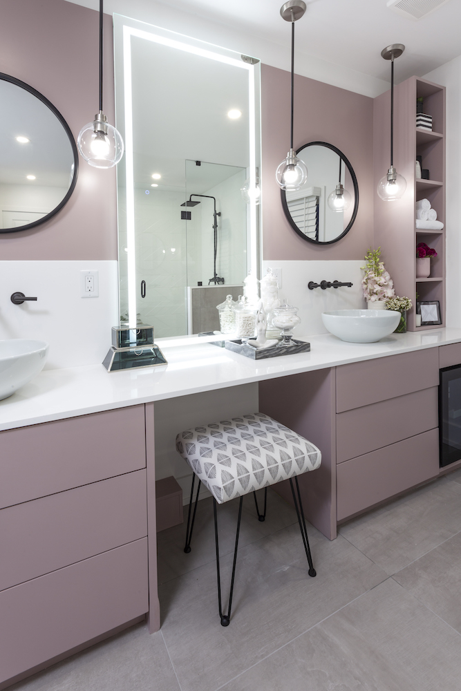 Two round mirrors and pink bathroom cabinets with a grey and white patterned stool tucked under a white vanity