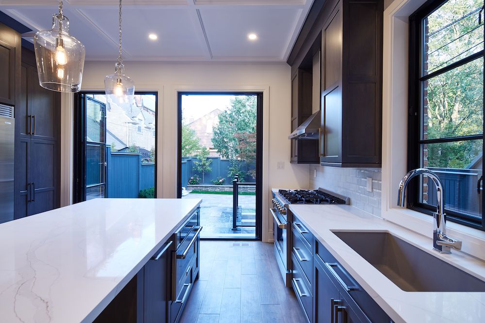 View of a modern kitchen with large centre island, two glass pendant lights, white quartz countertops, dark cabinets, and white walls, looking out through a set of glass and metal doors made by Magic™ Windows