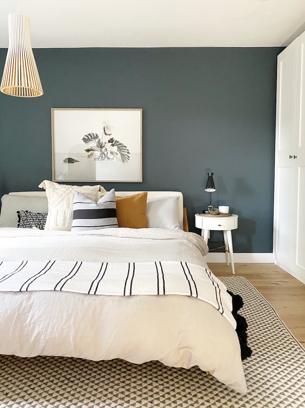 Beautiful bedroom designed and styled by The Property Stylist Inc. with a grey green painted feature wall behind a large white bed stacked with pillows, large pendant lamp, a geometric print rug and wood floors
