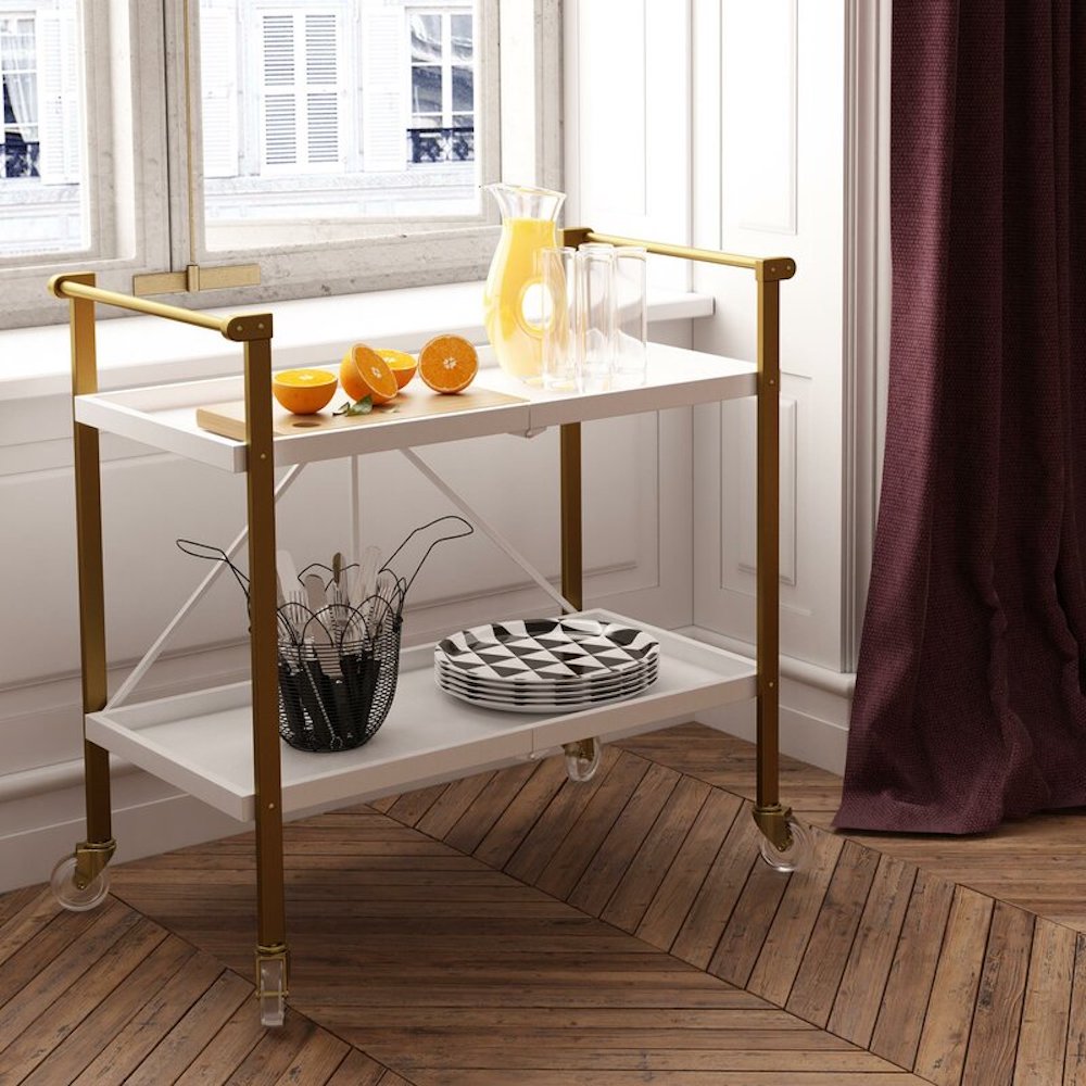 Gold and white bar cart with orange halves, a rug of orange juice, three glasses, a black basket filled with cutlery, and stack of black and white plates sitting on a herringbone wood floor under a white window