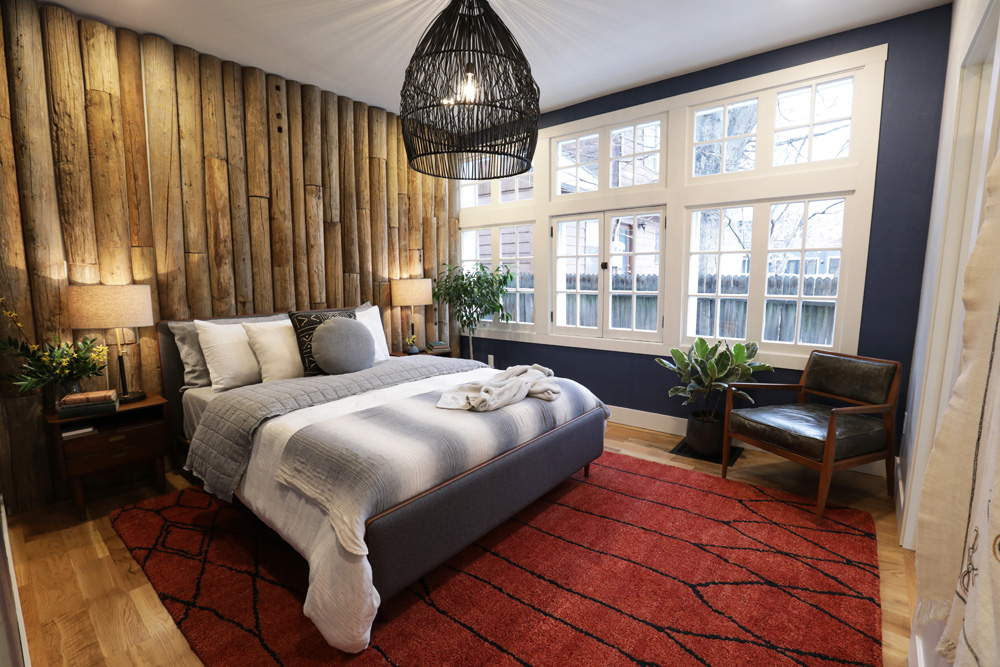Cozy cabin themed bedroom with a log feature wall behind the grey upholstered bed, large white framed windows, navy walls, a large red run, two bedside tables with laps, a large black wicker pendant lamp, and black leather and wood armchair in the corner designed by Alison Victoria on HGTV’s Rock the Block