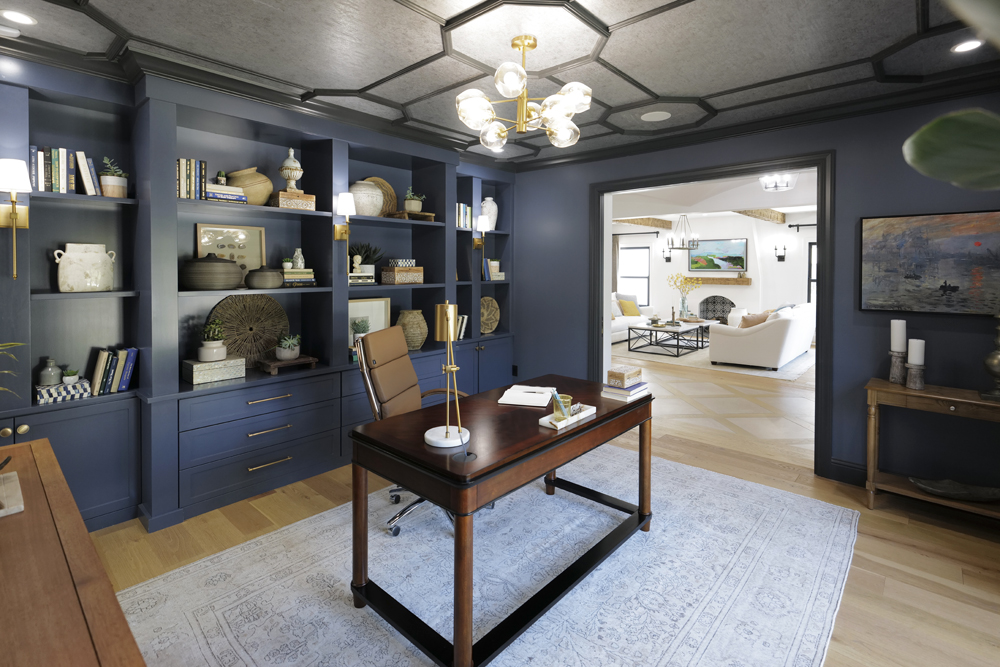 Sophisticated dark blue home office with black trim and ceiling moulding with built-in shelves and drawers with brass hardware, light wood floors, a dark wood desk and brown office chair as featured on HGTV’s Brother vs. Brother