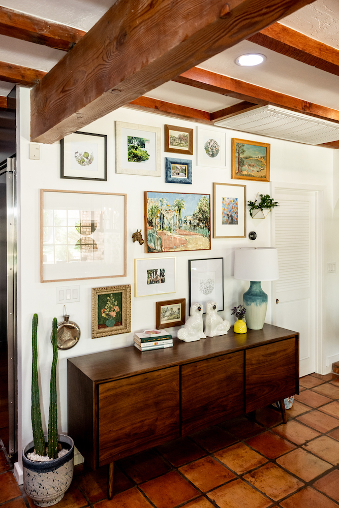 An eclectic art wall with framed paintings, a small bronze horse head and a succulent planter hung above a mid century modern credenza