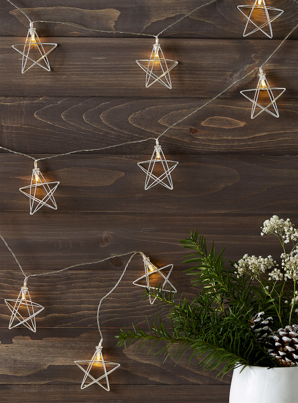 Silver star string lights from Simons hanging on a wood board wall with a white vase filled fir branches, pinecones and white flowers