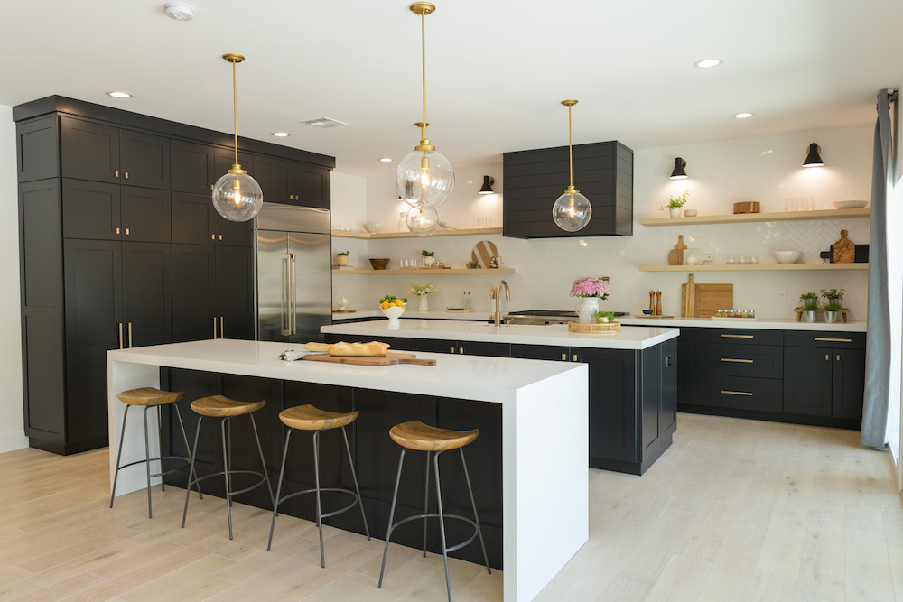 Sophisticated kitchen designed by Jonathan and Drew Scott for a remodel on HGTV’s Property Brothers with black floor to ceiling cabinets, gold hardware, white countertops and three glass globe pendant lights