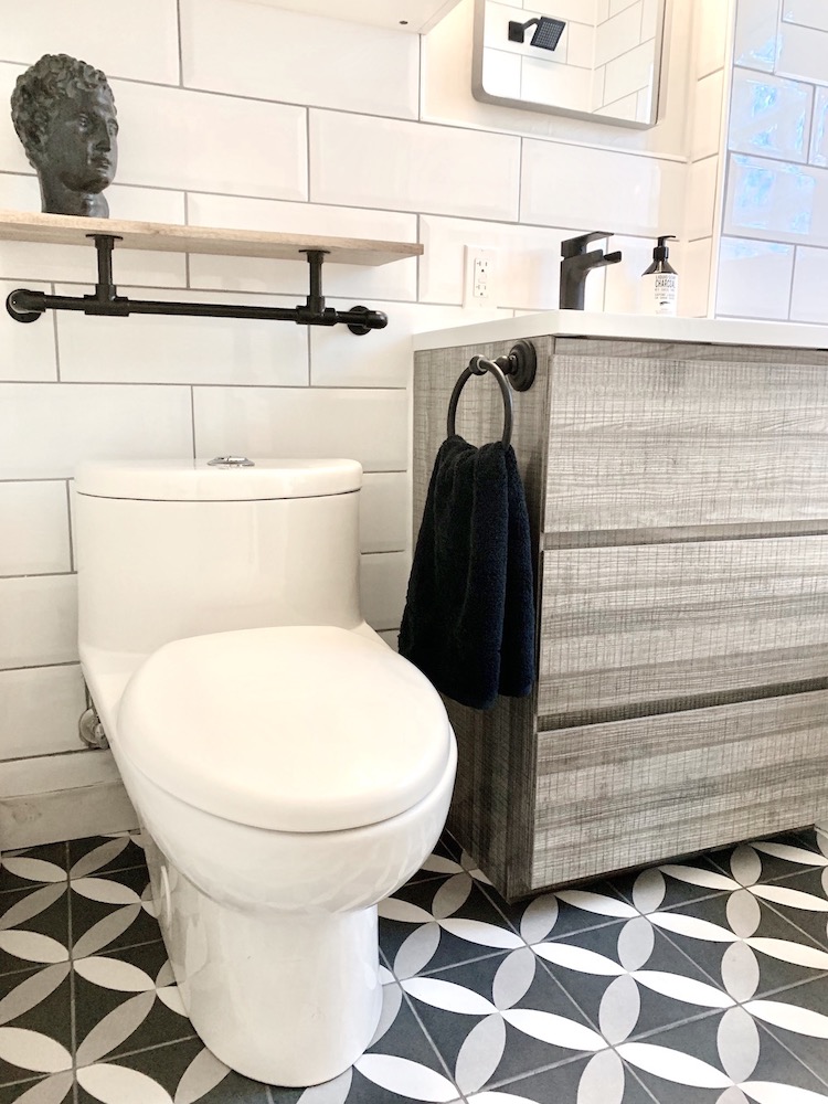 Beautiful modern bathroom with white and grey patterned floor tiles, white toilet, grey vanity with a white sink and a black faucet, grey mirror, and a floating industrial shelf with a black bust