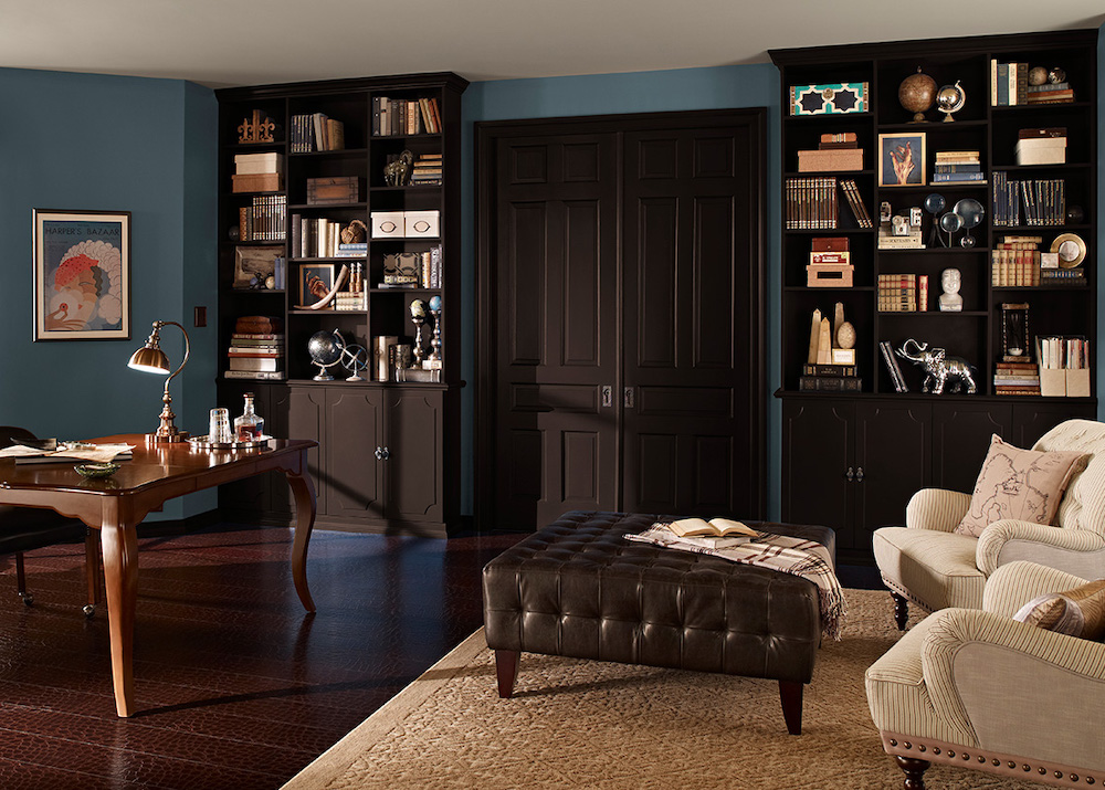 Stately library with two built in shelves full of books, a desk, a brown leather ottoman, two beige armchairs, a beige rug, dark wood floors, and walls painted in BEHR Blueprint S470-5