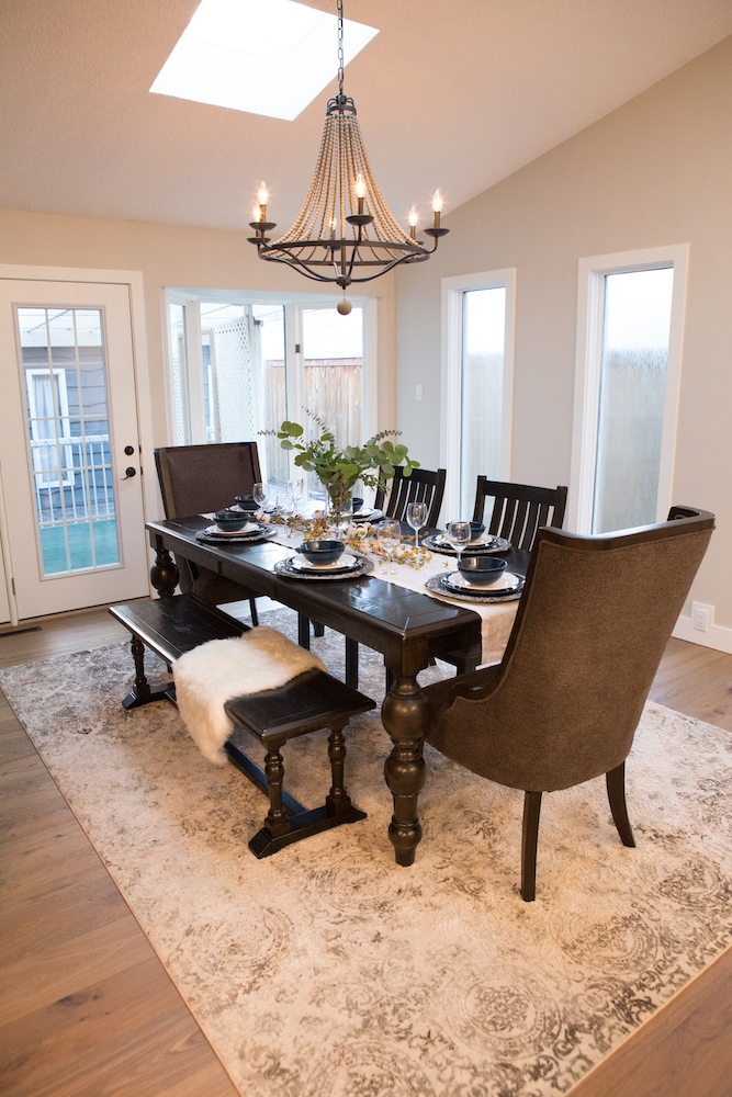 Light and airy traditional dining room featured on The Property Brothers on HGTV with many tall windows, a large dark wooden table, a wooded bench with a white sheepskin and four other chairs, a cream coloured patterned rug, and a vintage inspired chandelier